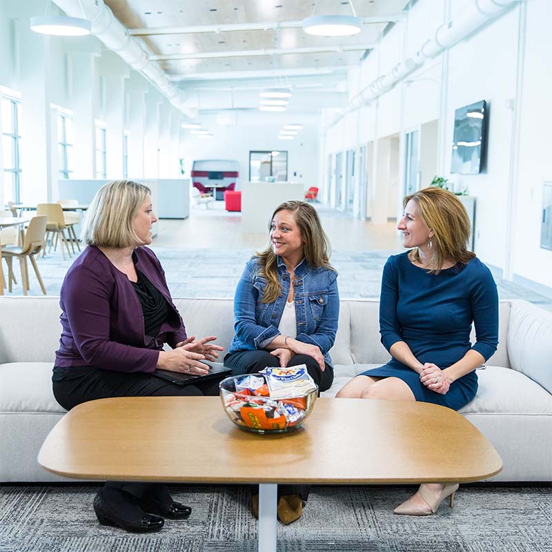 Three women sit together on a light-colored couch in a spacious, modern office, engaged in conversation. A wooden coffee table in front of them holds a glass bowl filled with assorted snacks, including candy bars and chips. The environment is bright with large windows, white walls, and contemporary furniture, creating a welcoming and collaborative atmosphere. The mood is friendly and professional.