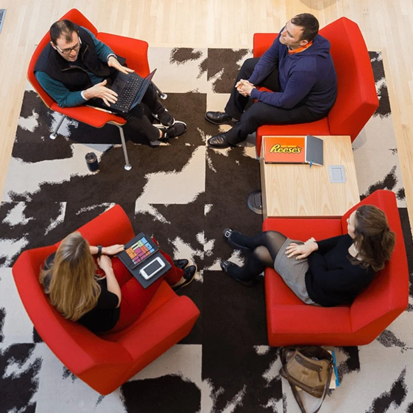 Four people sit in red armchairs arranged in a circle on a black and white patterned rug, engaged in a collaborative meeting. One person types on a laptop, another holds a colorful notebook, and a Reese's candy bar and notebook rest on a small wooden table. The setting is a modern, open office with light wood floors and a welcoming, focused atmosphere. No visible text appears in the image. The mood is professional and collegial, emphasizing teamwork and communication.