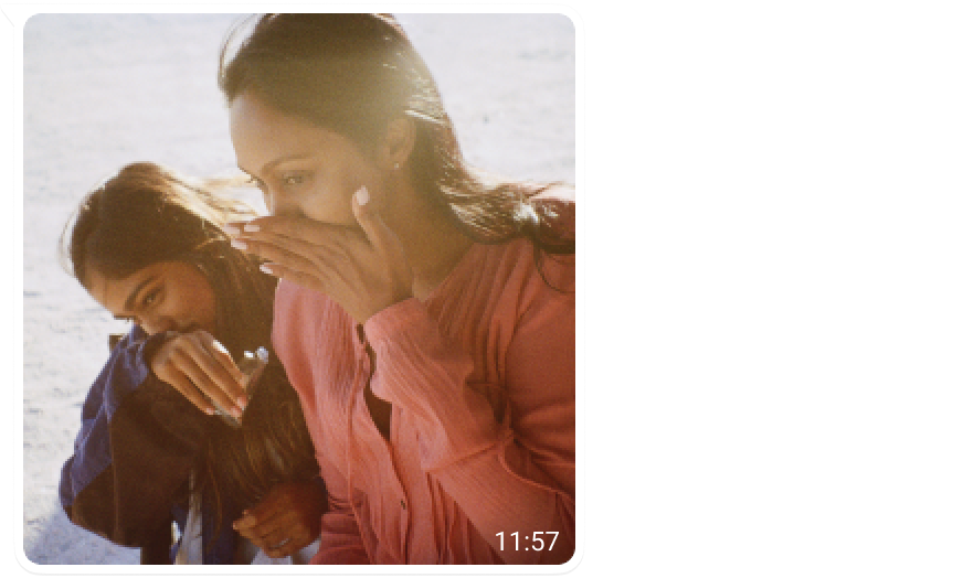 Two people standing close together outdoors, one wearing a pink shirt and the other in a blue jacket, both smiling and appearing joyful in bright natural light. The background is softly lit, suggesting a peaceful and warm atmosphere. No visible text is present in the image.