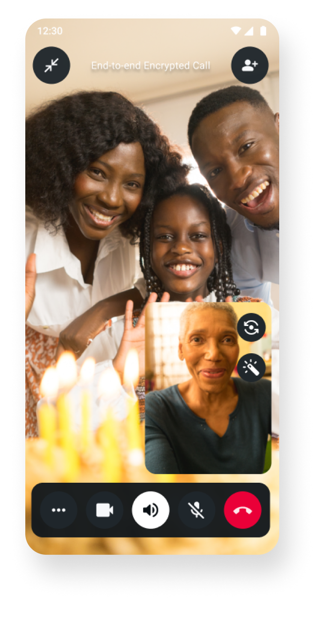 Video call interface showing three people gathered together smiling and waving, with a fourth participant in a smaller window at the bottom right. The background suggests a warm indoor setting with birthday candles in the foreground, creating a joyful and celebratory atmosphere. On-screen icons indicate call controls and end-to-end encrypted call status. The time 12:30 is displayed at the top.